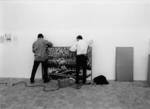 Zwei Männer, in Rückenansicht zu sehen, bereiten das aufgezogene Foto für die Performance vor.