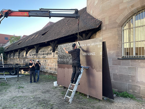 Documentation of the performance. A posterer attaches the print to the steel plate. He is standing on a short ladder and a crane can be seen in the background.