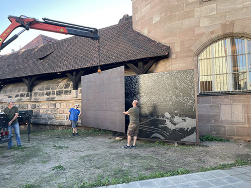Using a crane controlled by a person on the left of the image, a second steel plate is swung in from the left in front of the first steel plate with the image. Two assistants align the plate.