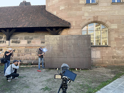 A worker wearing face protection welds the edges of the two steel plates. Two photographers and a centrally positioned tripod with a camera can be seen.